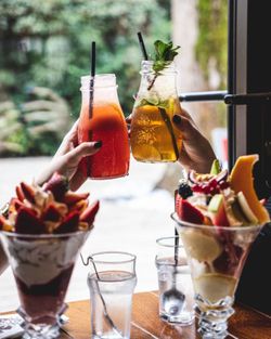 Deux cocktails colorés avec pailles tenus au-dessus de deux coupes de dessert aux fruits sur une table en bois.