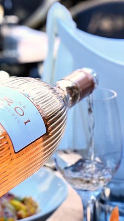 Bouteille de vin rosé Le Roi Soleil versée dans un verre, sur une table avec assiette et décor de terrasse.