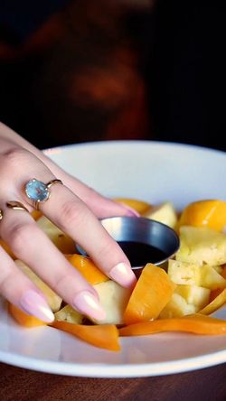 Assiette de fruits frais avec une main féminine portant une bague touchant les morceaux de fruits et un petit bol de sauce.