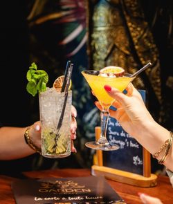 Deux mains tenant des cocktails variés, un mojito et un cocktail tropical, sur une table avec une ardoise en arrière-plan.