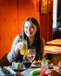 Jeune femme souriante dégustant une boisson sur la terrasse d'une brasserie avec plats et verres de vin.