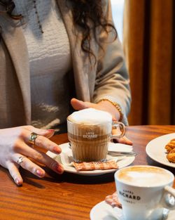 Tasse de café mousseux servie sur table en bois dans une brasserie avec une assiette de croissants à côté.