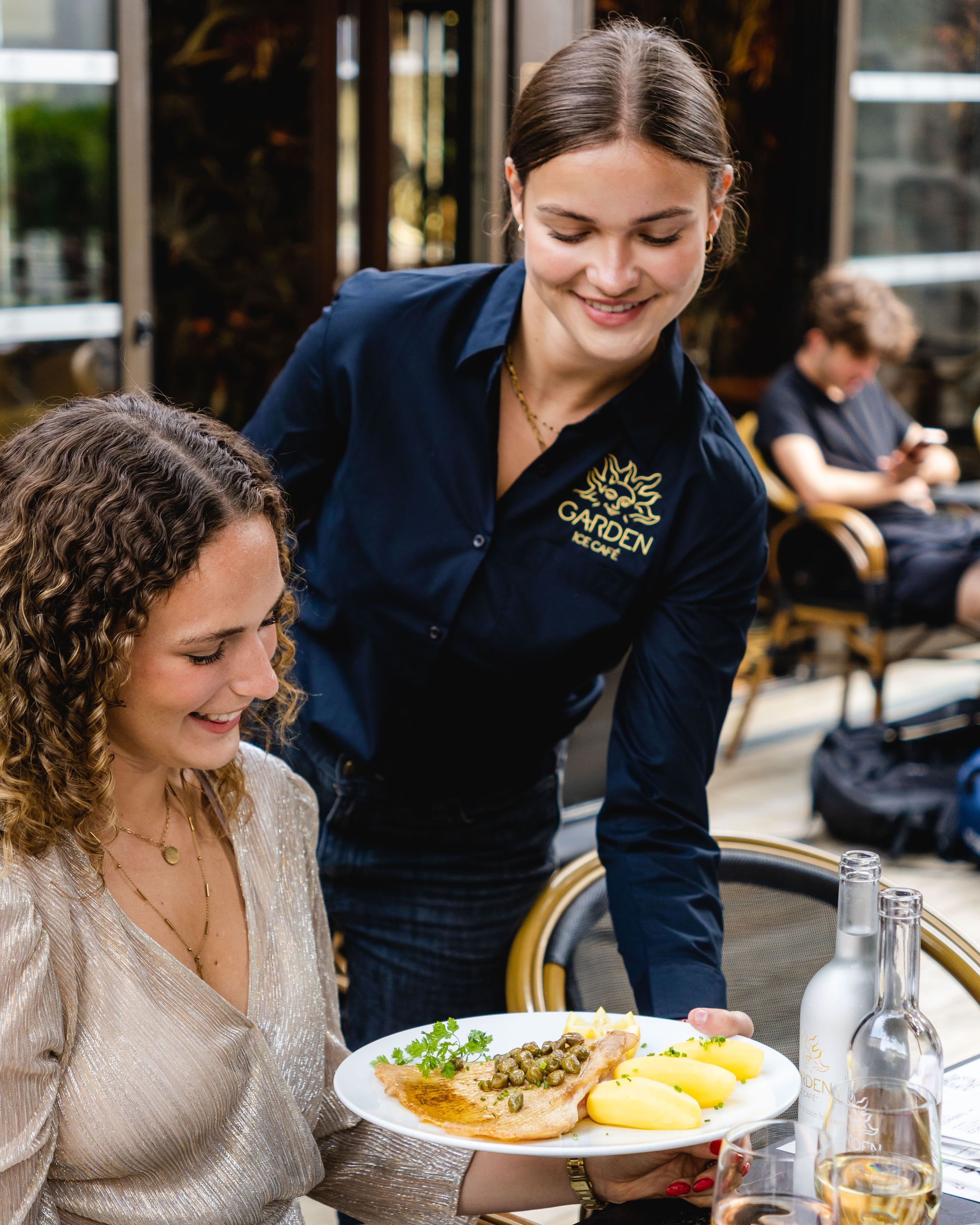 Serveuse du Garden Ice Café Périgueux servant un plat devant une cliente souriante en terrasse de brasserie.