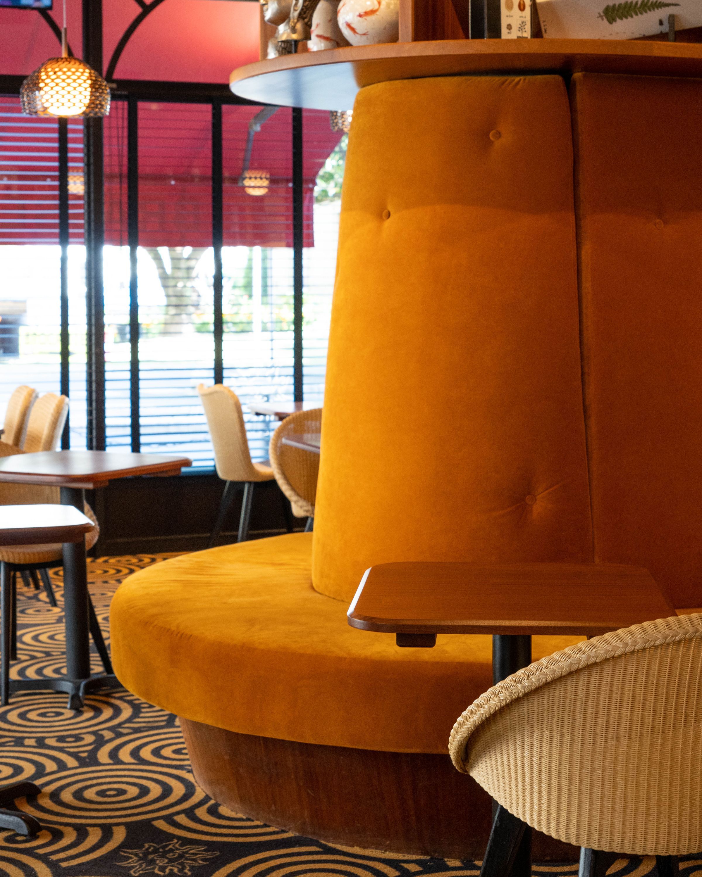 Espace intérieur d'un café avec banquette en velours orange et tables en bois près de grandes fenêtres.